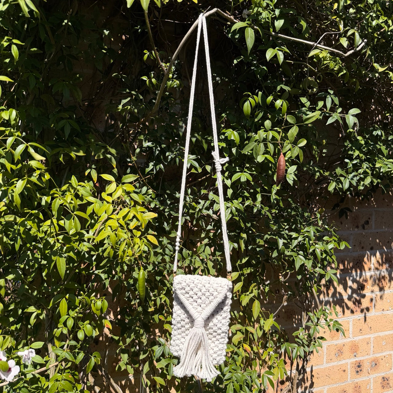 White macrame bag hanging from a branch with green leaves and a brick wall in the background