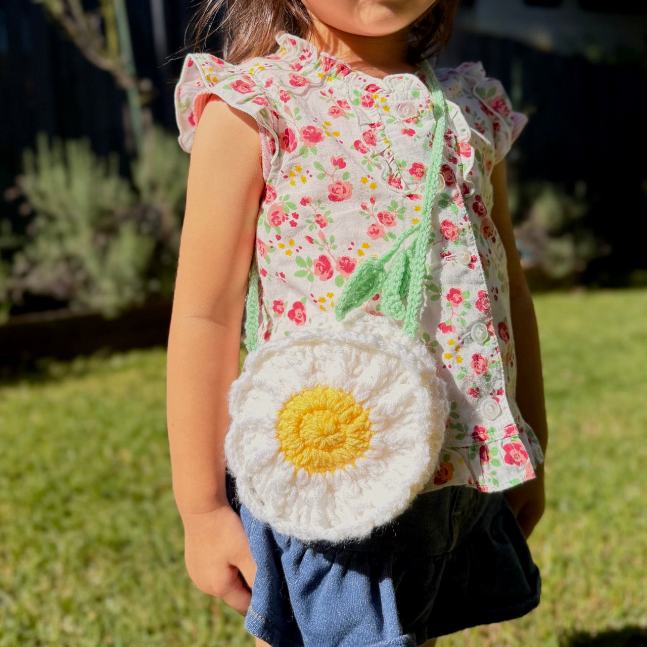 Child wearing a floral dress and holding a crochet bag with a yellow center outdoors.