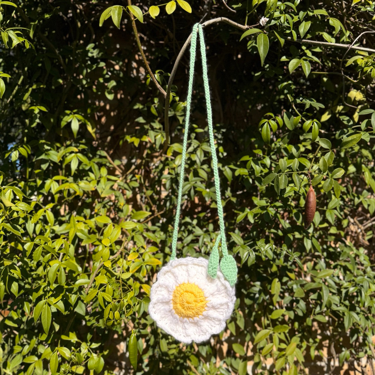 Crocheted flower-shaped bag with a yellow center and white petals hanging from green string against a leafy green background.